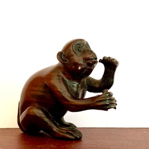 May include: A dark brown wooden monkey figurine sitting with one hand raised to its ear. The monkey is carved in a realistic style with detailed features.