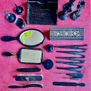 Könnte beinhalten: Ein Vintage-Schminkset in Schwarz mit einem Handspiegel, einem kleinen rechteckigen Spiegel, einer Bürste, einem Kamm, einer Pinzette und anderen Werkzeugen. Das Set ist auf einer rosa Stoffoberfläche angeordnet.