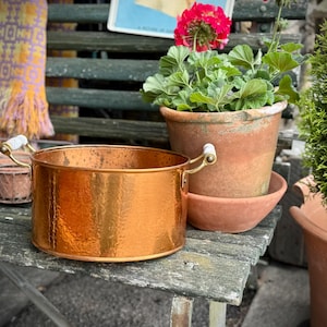 May include: A copper planter with a hammered finish and white and gold handles. It is placed on a weathered wooden surface, beside a terracotta pot with green plants and a red flower. Small terracotta pots are in a wire basket.