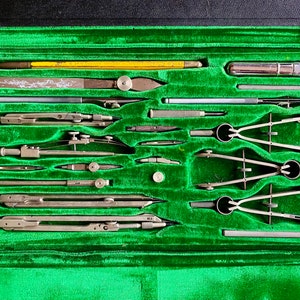 Può includere: Un set da disegno vintage in una custodia nera con interno in velluto verde. Il set include vari strumenti da disegno in metallo, come compassi, divisori e righelli. È visibile anche una matita gialla. La custodia è aperta, mostrando gli strumenti ben disposti.