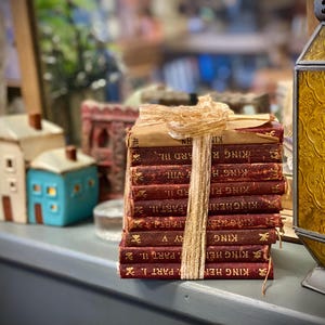 Puede incluir: Una pila de libros antiguos de tapa dura roja con letras doradas. Los libros están atados con una cinta beige y los títulos incluyen "King Henry", "King Richard" y "King John".