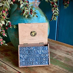 May include: An open wooden box containing metal letterpress type, with a decorative emblem on the lid. The box sits on a distressed wooden surface, with a large turquoise vase and greenery in the background.