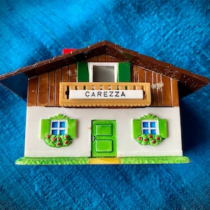 Può includere: Una piccola casa di legno bianca e marrone con tetto verde e persiane verdi. La casa ha un balcone con un cartello che dice "Carezza".