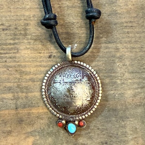 Pode incluir: Um colar com pingente de prata com um cordão de couro preto. O pingente tem um design circular com padrões intrincados e é adornado com pedras vermelhas e turquesas. O colar é exibido em uma superfície de madeira.