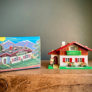 May include: A small, white, and red model house with a green balcony and red shutters. The house is sitting on a wooden surface with a box behind it. The box is blue and has a picture of a house on it.