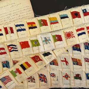Peut inclure: Collection de cartes de cigarettes anciennes représentant des drapeaux de différents pays. Les cartes sont disposées en bandes, avec le texte "American Cigarettes" et "British Empire". Un document manuscrit est visible dans le coin supérieur gauche.