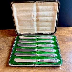 May include: A set of six silver-plated butter knives with ornate handles, presented in a black rectangular case with a green velvet interior. The interior lid features the text Mappin & Webb Ltd, London, Sheffield, England.