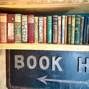 May include: A wooden shelf holds a collection of antique books with varied spine colours. The books have visible titles and decorative details. Below, a sign reads "BOOK HERE" with an arrow pointing left.
