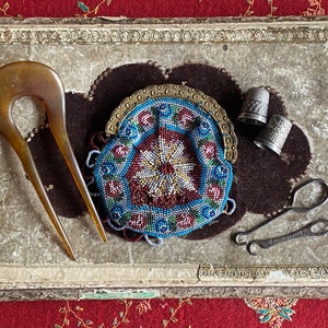 Puede incluir: Un kit de costura vintage con una bolsa de cuentas, dos dedal y un par de enhebradores de agujas. El kit se muestra en una cubierta de libro antiguo desgastada.