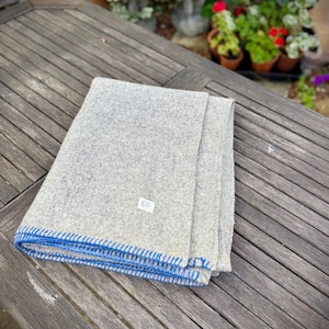 May include: A folded grey blanket with blue stitching along the edges. The blanket is made of a soft, textured material and is folded neatly on a wooden table. A small white label is visible on the blanket.