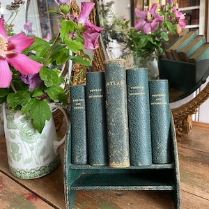 Puede incluir: Un conjunto de seis libros antiguos encuadernados en cuero verde con letras doradas en los lomos. Los libros se titulan "Poesía y prosa", "Diccionario alemán", "Atlas", "Diccionario francés", "Proverbios y mitología". Los libros se muestran en un expositor de libros de cuero verde con un estante.