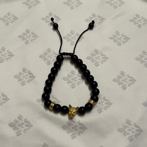 May include: A black beaded bracelet with a gold wolf head charm. The bracelet has a simple knot closure.