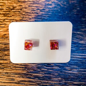 May include: A pair of square-shaped stud earrings with a pink, orange, and gold glitter embedded in clear resin.