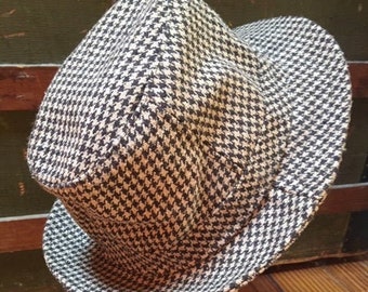 May include: A black and white houndstooth patterned hat with a brim. The hat is sitting on a wooden surface.