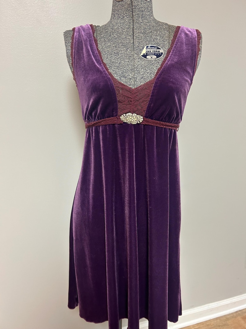 Op de afbeelding: Een mouwloze, knielange paarse fluwelen jurk. De jurk heeft een V-hals met bordeauxrode kanten afwerking en een bordeauxrode empire taille. Een decoratieve strass broche accentueert de taille. De jurk wordt op een mannequin getoond.