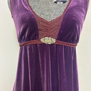 Op de afbeelding: Mouwloze, vloerlange paarse fluwelen jurk met een V-hals. Het lijfje heeft een bordeaux kanten inzetstuk en een decoratieve strass sluiting in de taille. De jurk wordt gepresenteerd op een mannequin.