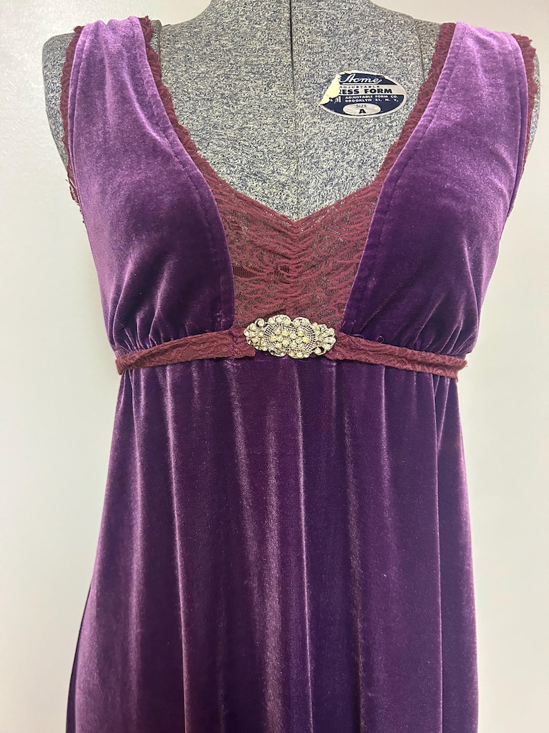 Op de afbeelding: Mouwloze, vloerlange paarse fluwelen jurk met een V-hals. Het lijfje heeft een bordeaux kanten inzetstuk en een decoratieve strass sluiting in de taille. De jurk wordt gepresenteerd op een mannequin.