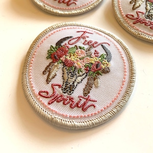 May include: A set of embroidered patches with a white background and a beige border. The patches feature a skull with flowers and feathers, and the text "Free Spirit".