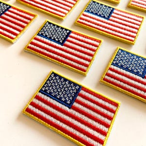 May include: Embroidered patches featuring the American flag. Each patch has a blue square with white stars in the upper left corner, red and white stripes, and a gold border. The patches are rectangular and are arranged on a white surface.