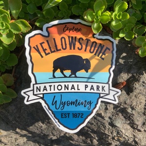 May include: A sticker featuring a design for Yellowstone National Park. It includes a bison silhouette, the text "Yellowstone National Park Wyoming" and "EST 1872". The background has orange, yellow, and blue gradients.