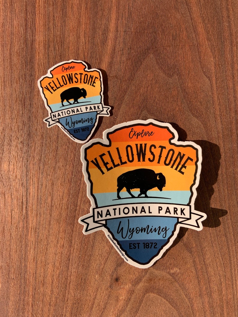 May include: Two Yellowstone National Park stickers. The stickers are shaped like an arrowhead and feature a bison silhouette, the words "Explore Yellowstone National Park Wyoming EST 1872", and a colourful sunset design.