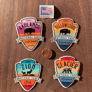 May include: Four colourful National Park stickers featuring illustrations of a bison, bear, and mountain goats. The stickers are for Badlands, Yellowstone, Zion, and Glacier National Parks. A Forever USA stamp and a penny are also in the image.