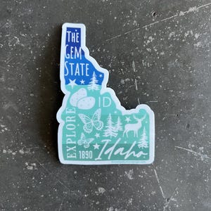 May include: A blue and green sticker shaped like the state of Idaho. It features the text "The Gem State" and "Explore Idaho 1890" along with images of butterflies, trees, potatoes, and a deer.