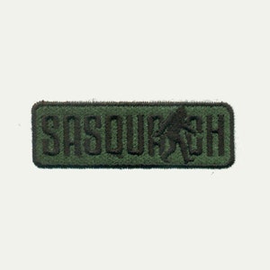 Pode incluir: Etiqueta bordada verde com a palavra "SASQUATCH" em letras pretas e uma silhueta preta de um pé-grande caminhando.