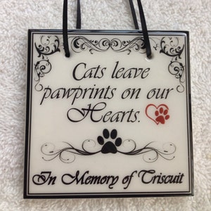 May include: White ceramic tile with black trim and a black ribbon hanger. The tile has a black and white floral design with the text "Cats leave pawprints on our Hearts." A red heart with a black paw print is in the upper right corner. A black paw print is below the text. The bottom of the tile has the text "In Memory of Triscuit."