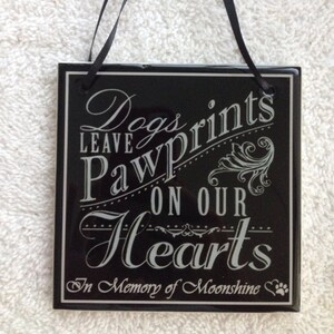 May include: Black ceramic plaque with white text that reads "Dogs leave pawprints on our hearts" and "In Memory of Moonshine". The plaque has a black ribbon for hanging.