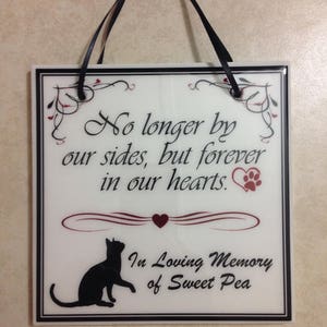 May include: A white ceramic plaque with black lettering and a black silhouette of a cat. The plaque reads "No longer by our sides, but forever in our hearts." with a red heart and paw print. Below the text is "In Loving Memory of Sweet Pea".