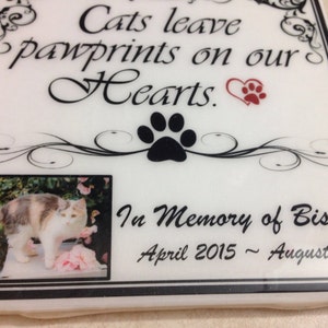 May include: White ceramic memorial plaque with black and white floral design. A black paw print is in the center. A photo of a calico cat is in the lower left corner. The text reads "Cats leave pawprints on our Hearts." and "In Memory of Biscuit April 2015 - August 2016".