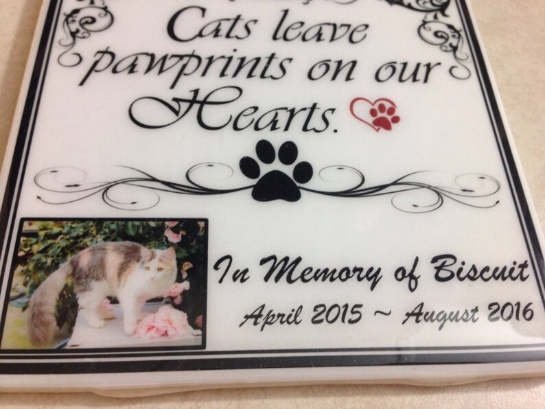 May include: White ceramic memorial plaque with black and white floral design. A black paw print is in the center. A photo of a calico cat is in the lower left corner. The text reads "Cats leave pawprints on our Hearts." and "In Memory of Biscuit April 2015 - August 2016".