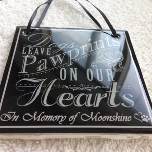 May include: Black ceramic tile with white text that reads "Dogs leave paw prints on our hearts. In memory of Moonshine."  The tile has a paw print and heart symbol.