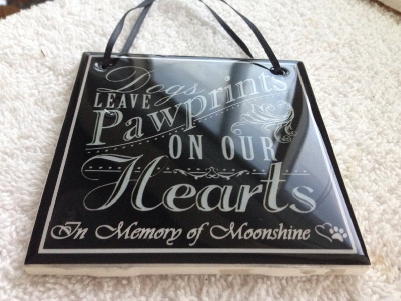 May include: Black ceramic tile with white text that reads "Dogs leave paw prints on our hearts. In memory of Moonshine."  The tile has a paw print and heart symbol.