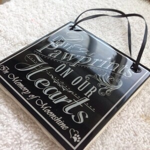 May include: Black ceramic tile with white text that reads "Leave Pawprints on our Hearts" and "In Memory of Moonshine". The tile has a black ribbon attached for hanging.