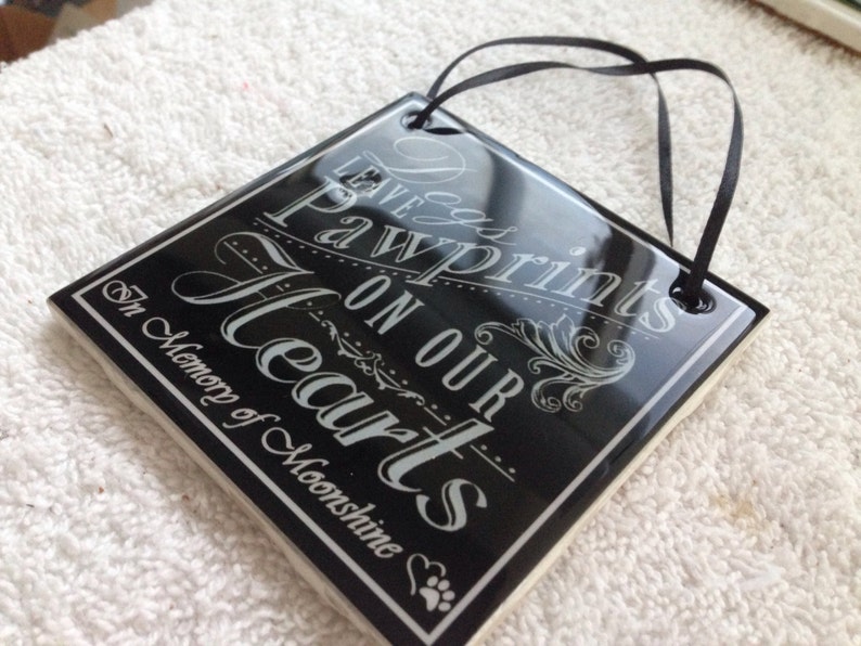 May include: Black ceramic tile with white text that reads "Leave Pawprints on our Hearts" and "In Memory of Moonshine". The tile has a black ribbon attached for hanging.
