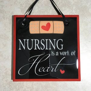 May include: Black and red square sign with a white bandage with a red heart on it. The sign reads "Nursing is a work of heart" with a small red heart at the end.