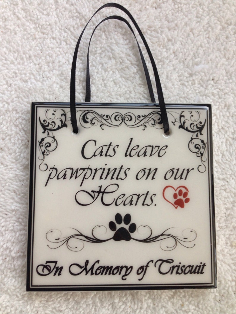 May include: White ceramic tile with black trim and a black ribbon hanger. The tile has a black and white floral design with the text "Cats leave pawprints on our Hearts." A red heart with a black paw print is in the upper right corner. A black paw print is below the text. The bottom of the tile has the text "In Memory of Triscuit."