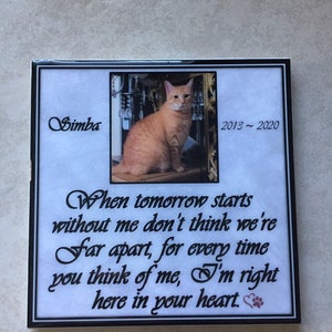 K&ouml;nnte beinhalten: Eine wei&szlig;e Keramikfliese mit schwarzem Rand und einem Foto einer orangefarbenen Tabby-Katze. Die Fliese tr&auml;gt ein Zitat, das lautet: "Wenn der Morgen ohne mich beginnt, denk nicht, dass wir weit entfernt sind, denn jedes Mal, wenn du an mich denkst, bin ich genau hier in deinem Herzen." Der Name der Katze ist Simba und die Daten 2013-2020 sind auf der Fliese aufgedruckt.