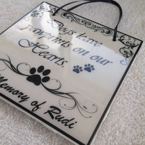 May include: White ceramic plaque with black lettering and a black paw print. The plaque reads "Dogs leave pawprints on our Hearts. In Memory of Rudi."