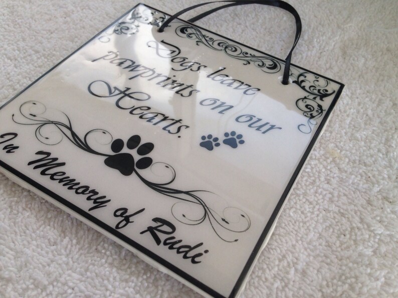 May include: White ceramic plaque with black lettering and a black paw print. The plaque reads "Dogs leave pawprints on our Hearts. In Memory of Rudi."