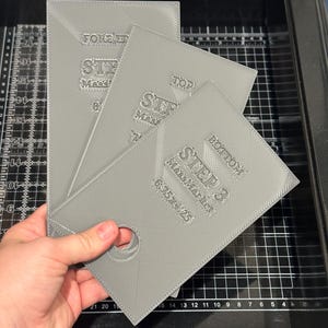 May include: A stack of three gray, rectangular plastic templates. Each template has the text "STEP 3 MassMarket 6.75x4.25" and other text. One template has a hole. The templates are held by a hand.