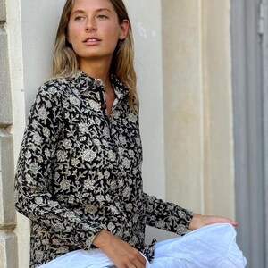 Puede incluir: Una camisa negra con estampado floral con mangas largas y una falda blanca escalonada. La camisa tiene un cuello alto y se combina con sandalias marrones. El atuendo es casual y elegante.