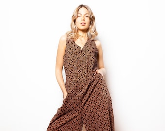 May include: A sleeveless, wide-leg jumpsuit in a brown and black geometric pattern. The jumpsuit has a V-neck and pockets. The model is wearing brown heeled sandals. The background is a white wall and a wooden floor.