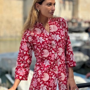 Puede incluir: Una camisa roja con estampado floral, mangas tres cuartos y botones frontales. La camisa presenta un patrón repetido de flores y hojas blancas y rosas. La modelo lleva pantalones blancos.