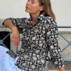 Puede incluir: Una camisa de manga larga con botones que presenta un fondo negro con un patrón floral blanco repetitivo. La camisa tiene un cuello clásico, una tapeta de botones en la parte delantera y puños en las mangas con un estampado botánico.