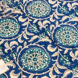 Può includere: Una tovaglia blu con un motivo floreale ripetuto in bianco e verde acqua. Il design presenta grandi fiori e foglie stilizzate, creando un tessuto vibrante e decorativo. Ideale per cene o occasioni speciali.