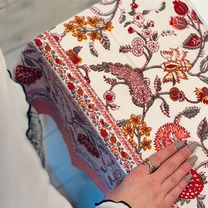 Puede incluir: Un mantel blanco con un estampado floral en rojo, amarillo y rosa. El diseño incluye flores y hojas estilizadas. El mantel tiene un borde rojo con un motivo floral repetido. El borde del mantel es rosa.