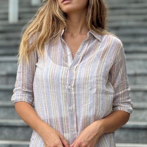 Può includere: Camicia abbottonata a righe multicolore con maniche arrotolate. La camicia presenta righe verticali nelle tonalità del blu, giallo e marrone. La modella indossa una gonna bianca. La camicia è in tessuto leggero.